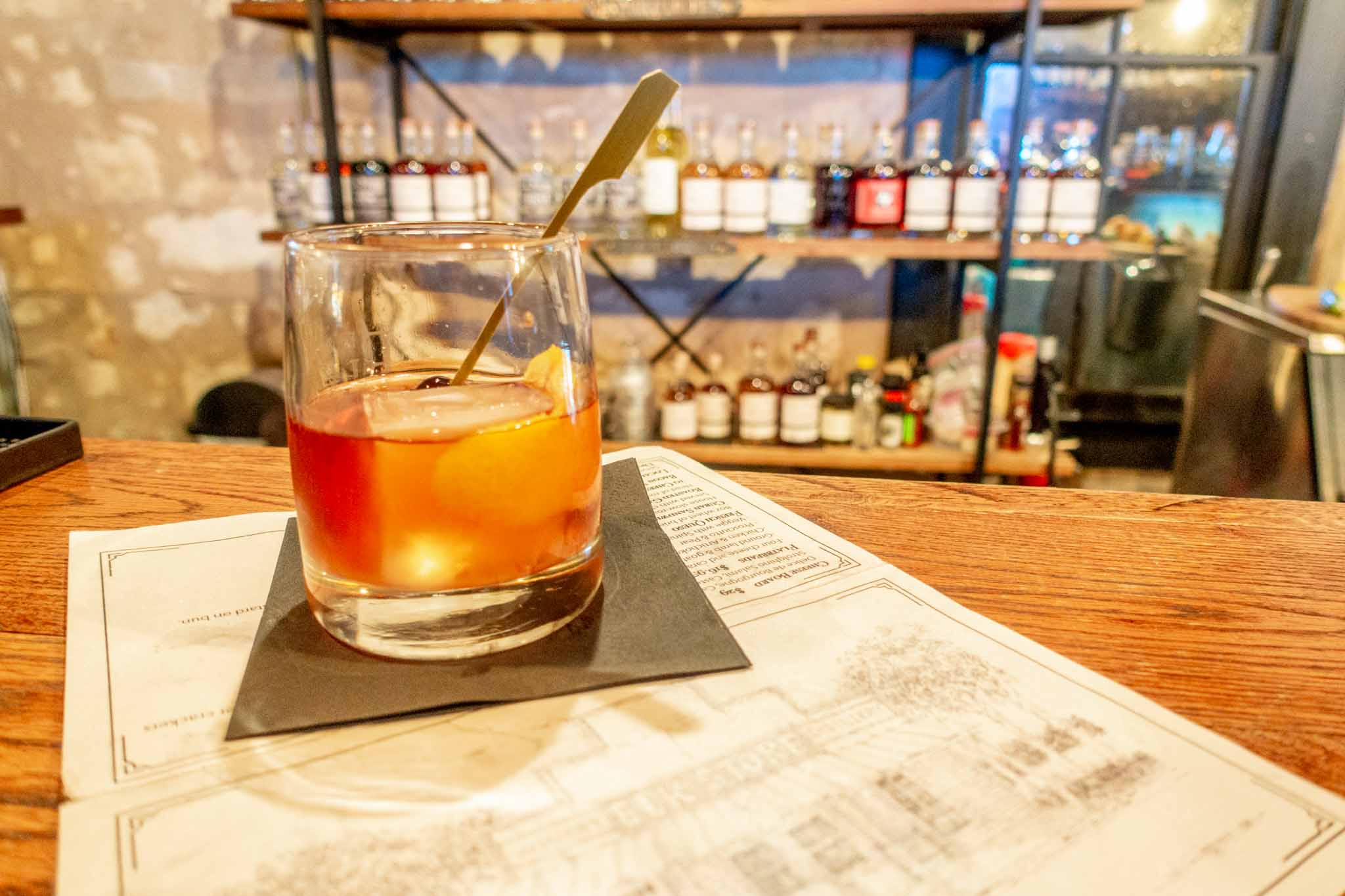 Whiskey cocktail on a bar in front of a shelf full of bottles.