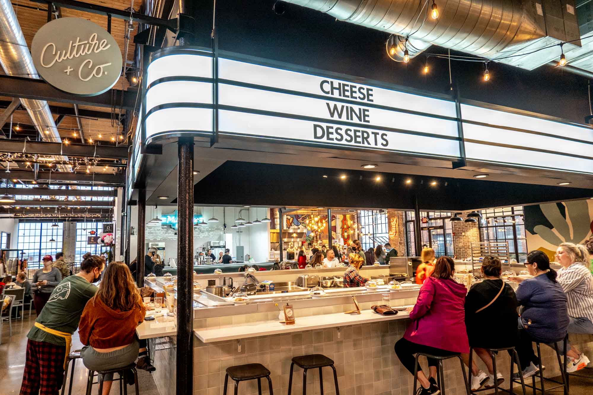 People sitting at a counter under a sign for "Culture + Co.: Cheese, Wine, Desserts."