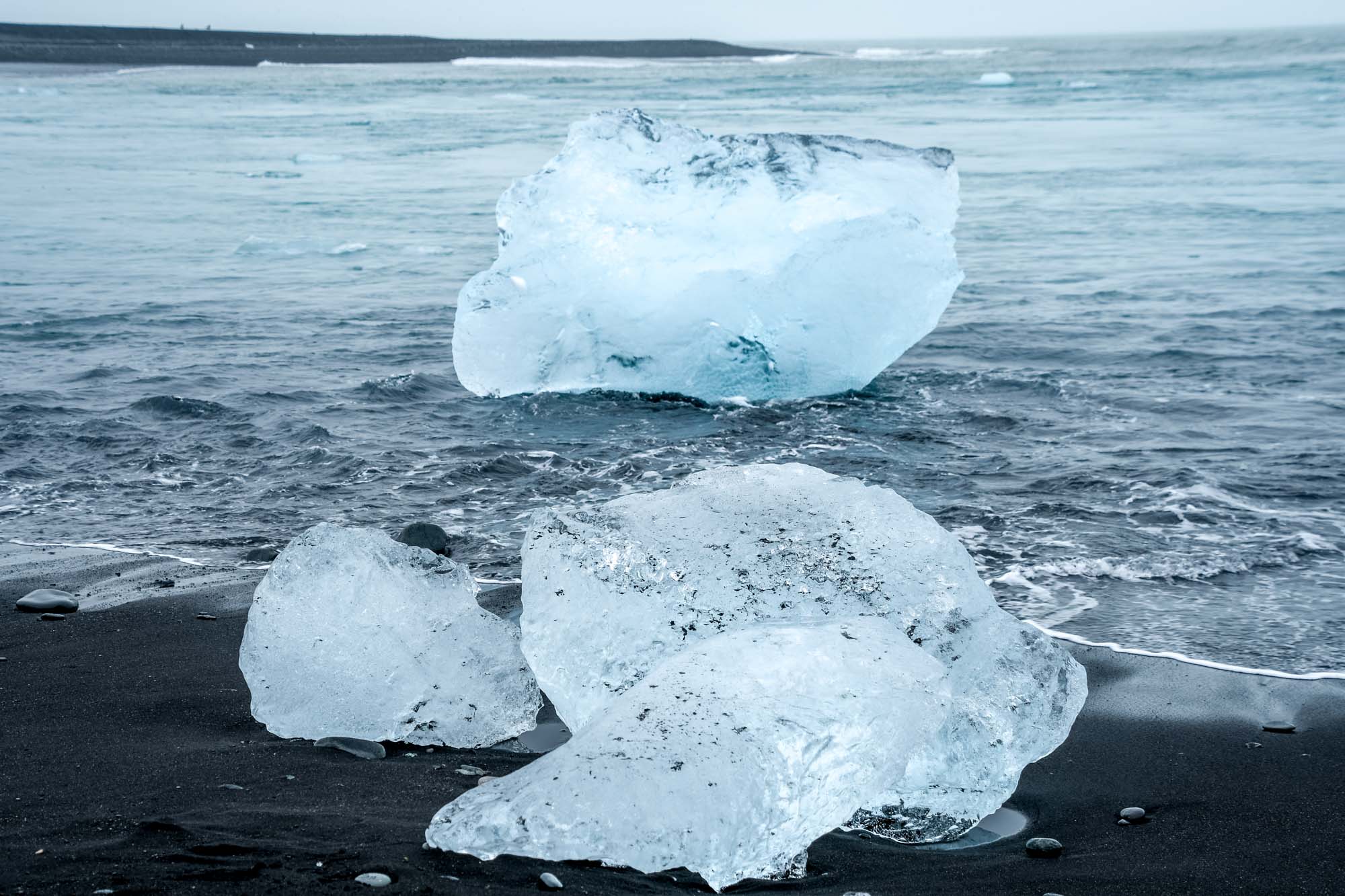 Large pieces of clear ice on a black sand beach being lapped by ocean waves