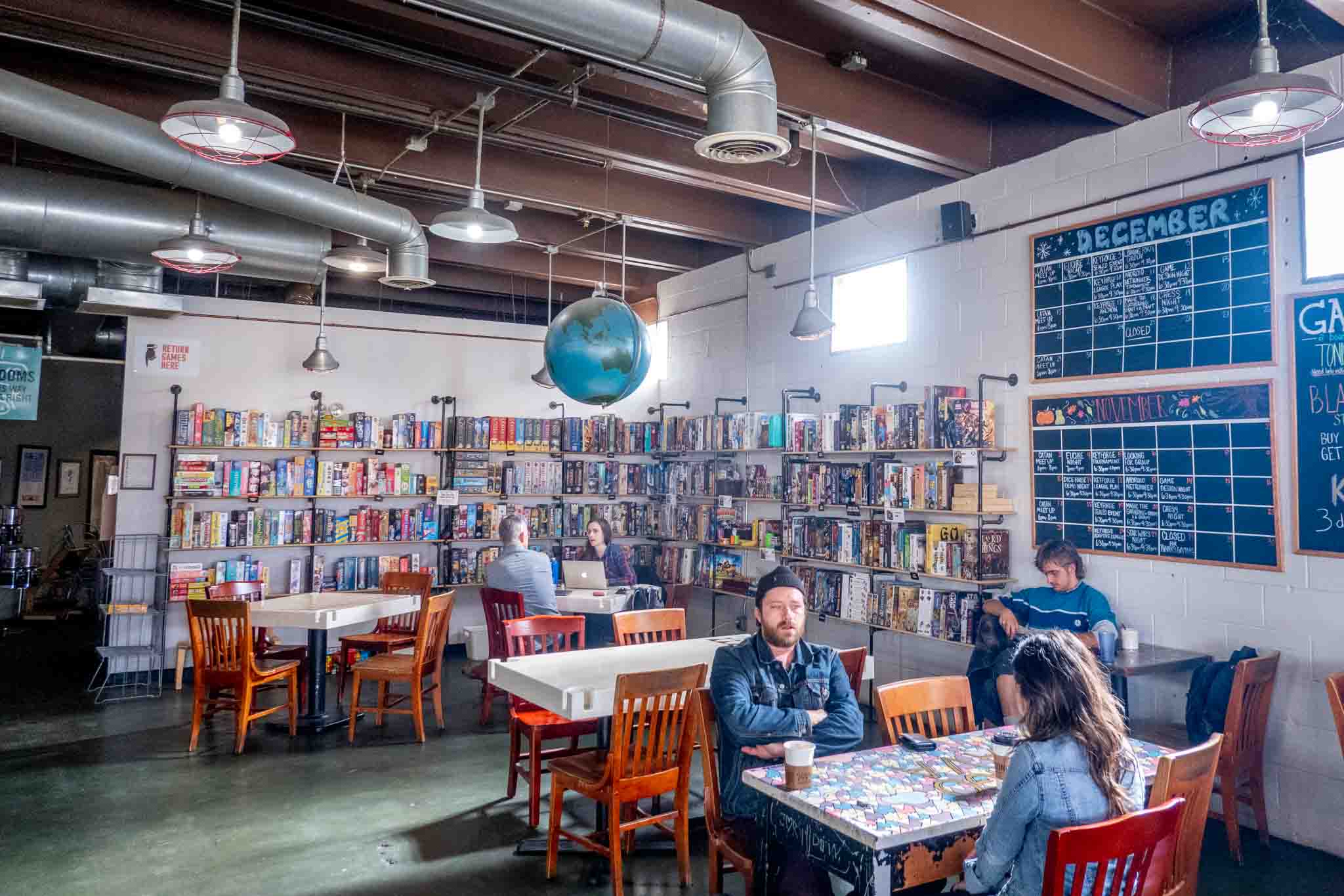 People at tables by a wall of board games. 