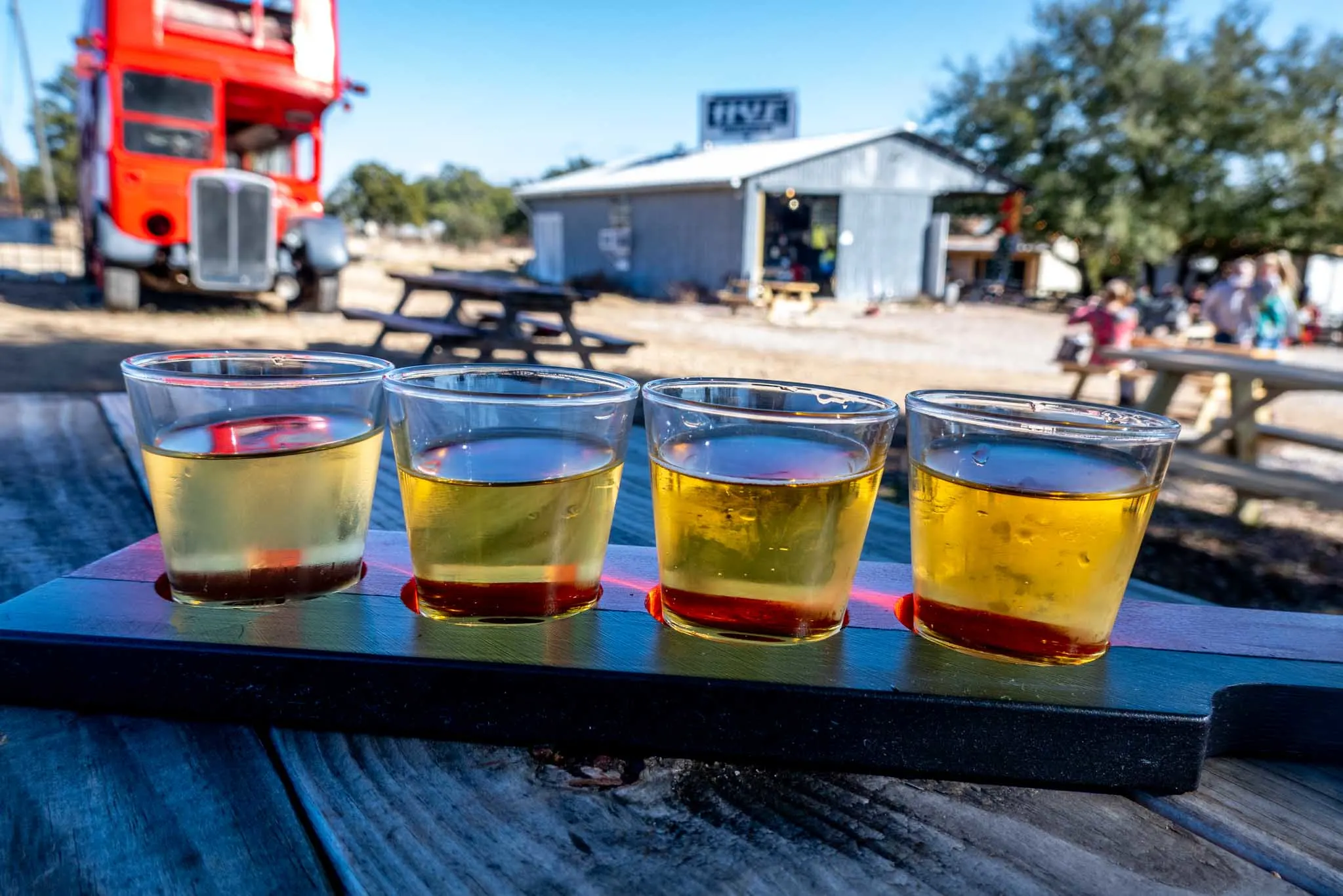 Flight of 4 glasses of cider on a picnic table.